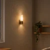 Kianna gold wall lamp providing warm illumination on a neutral wall beside decorative shelving